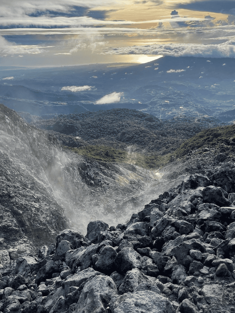 Steaming volcanic landscape with rocks and mountain views, cloudy sky, and sunlight in the background.