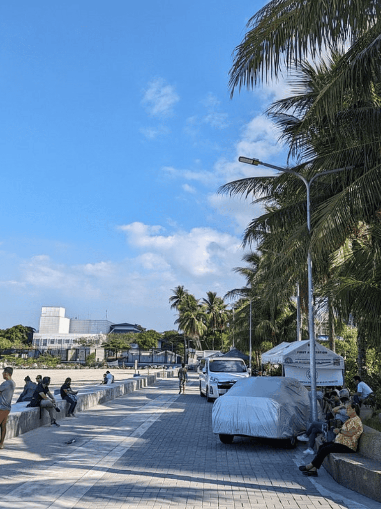 Bright coastal street with palm trees and people relaxing by the waterfront.