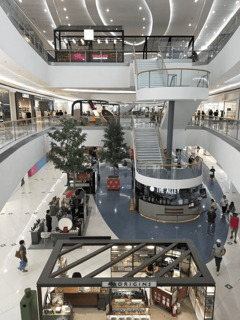 Bright modern shopping mall interior with multiple levels, store storefronts, and a central kiosk area.