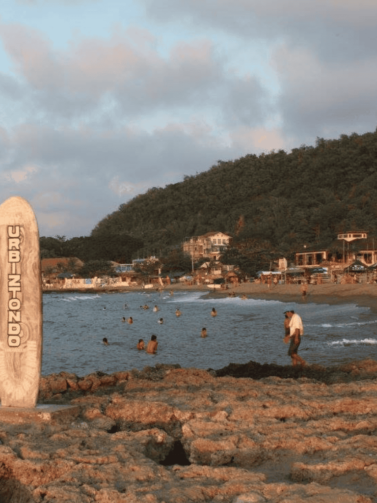 Beach with people swimming and relaxing, scenic coastal view, and lush green hills in the background.