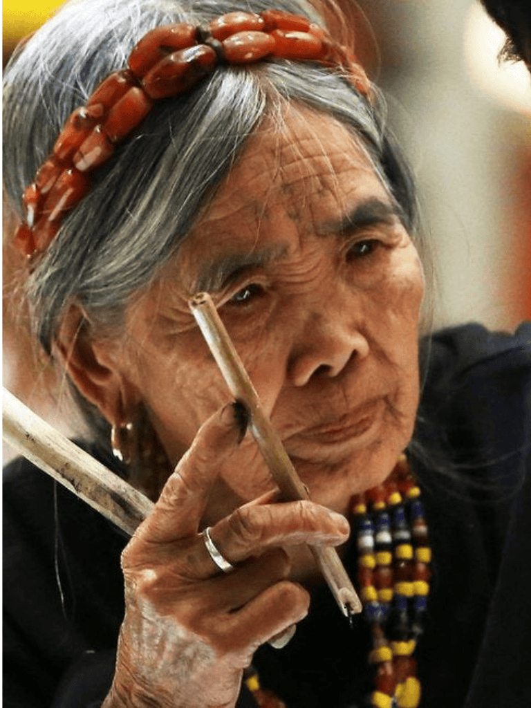 Wise elderly woman with traditional bead necklace and headband, holding a pen and studying intently.