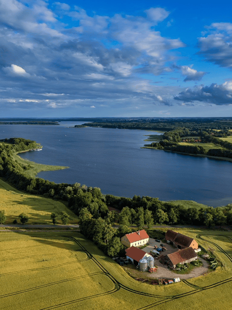 Serene lakeside landscape with farm buildings, lush greenery, and expansive sky, ideal for outdoor adventures and nature retreats.
