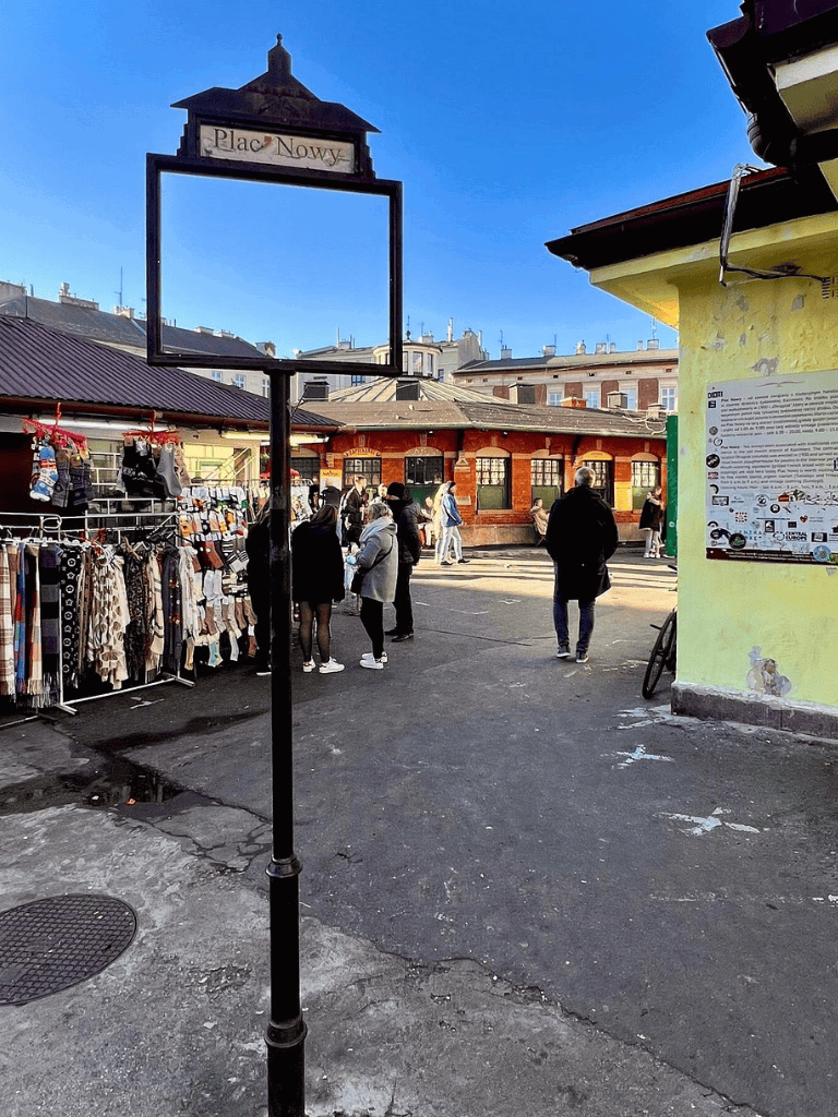 Busy outdoor market scene at Plac Nowy in Krakow, with shoppers and street vendors under clear blue skies.