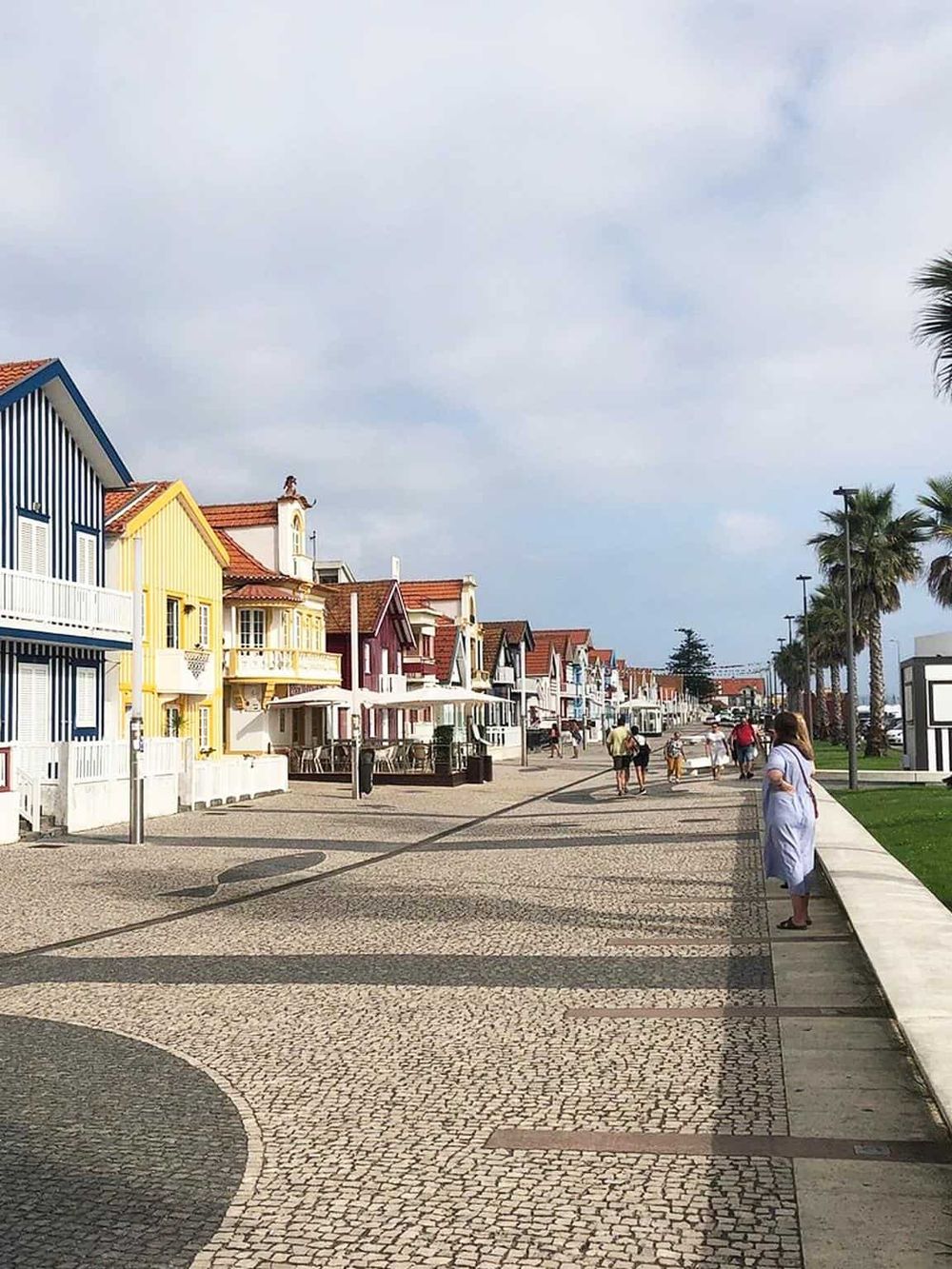 Colorful seaside houses along a pedestrian promenade with palm trees and relaxed visitors in the background.
