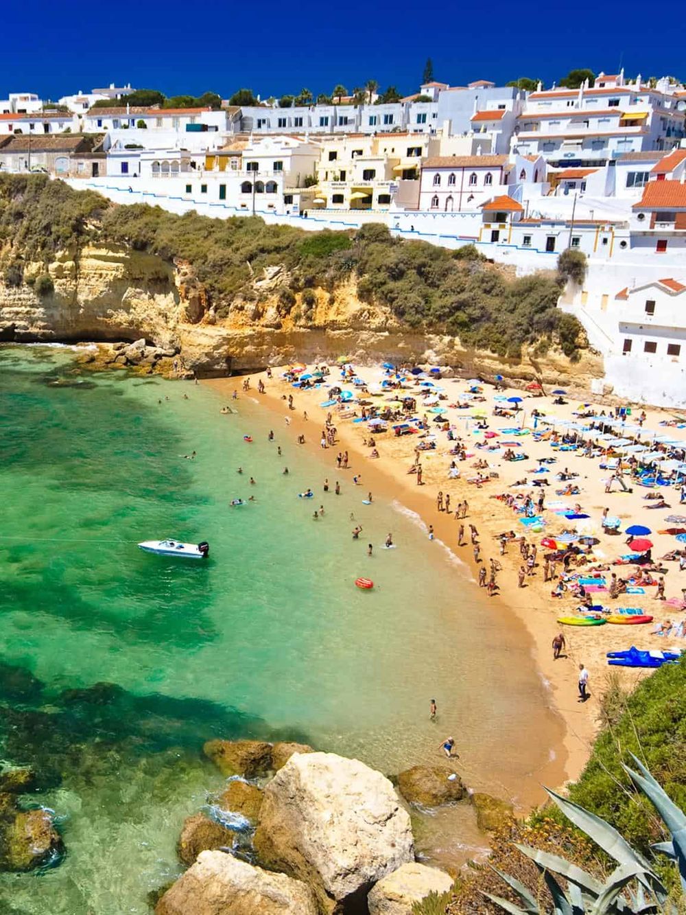 Vibrant beach scene with colorful umbrellas, turquoise waters, and hillside white houses in a coastal town.