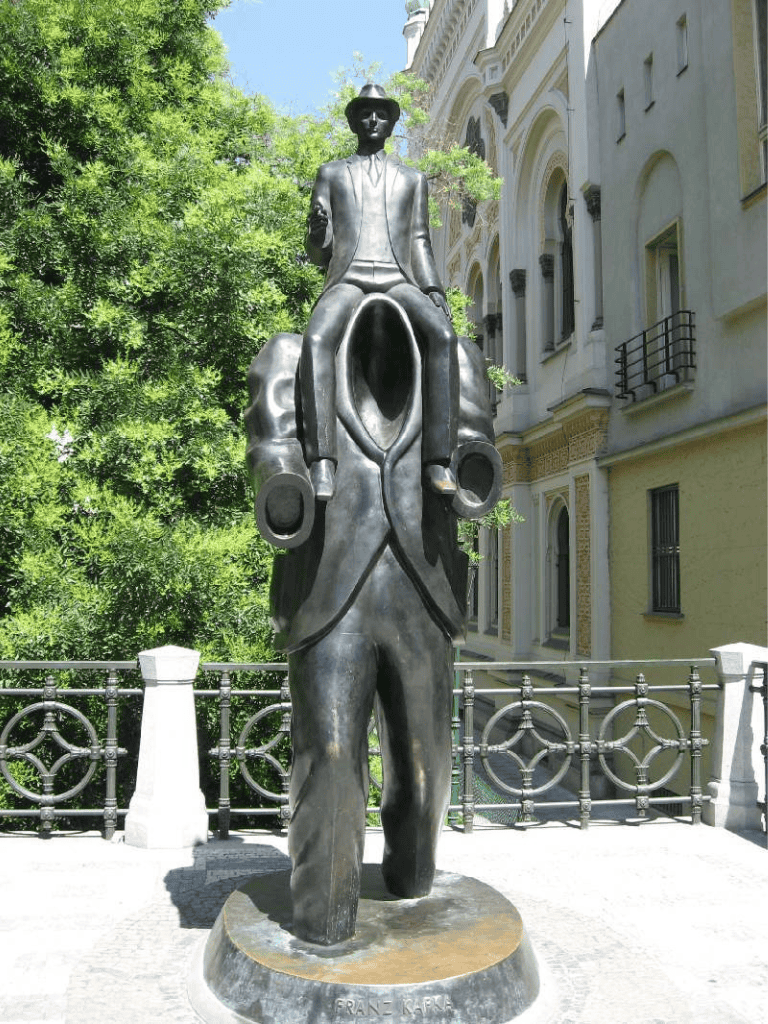 Sculpture of a man with a hat and suit, city street, green trees in background.