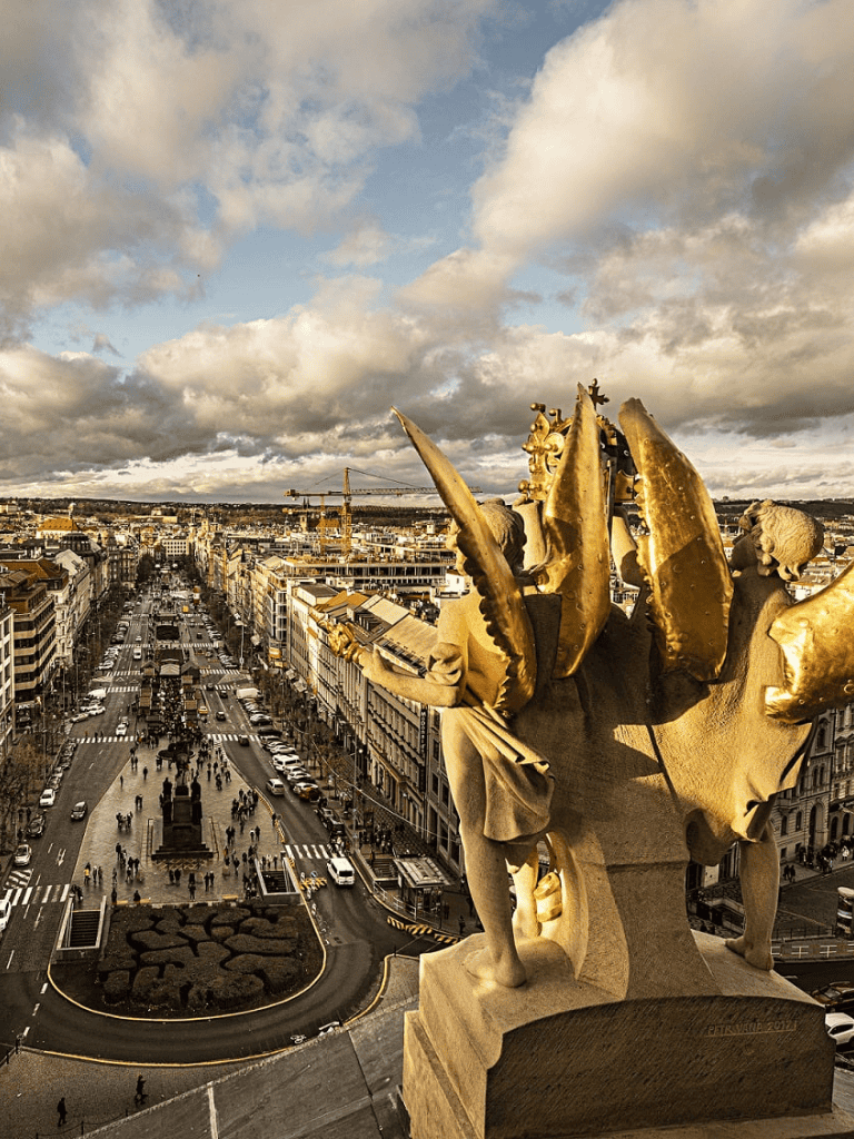 Intricate gold-winged statue overlooking a bustling city street in daytime.
