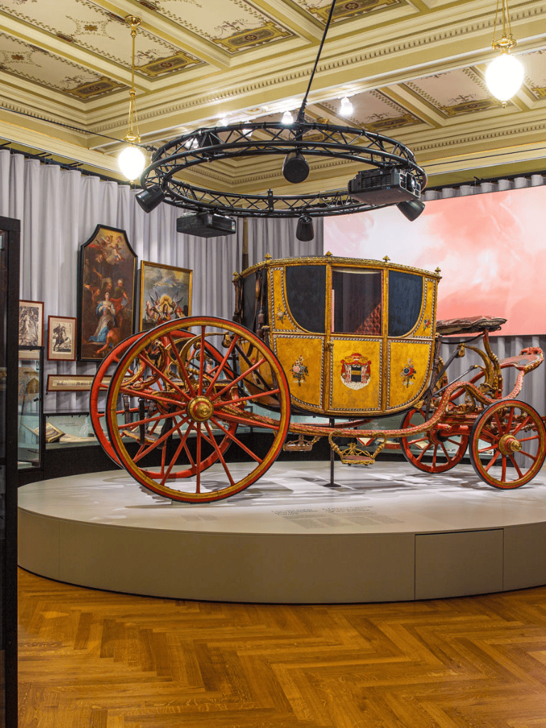 Ancient royal carriage in museum with paintings and ornate details, showcasing historical transportation.