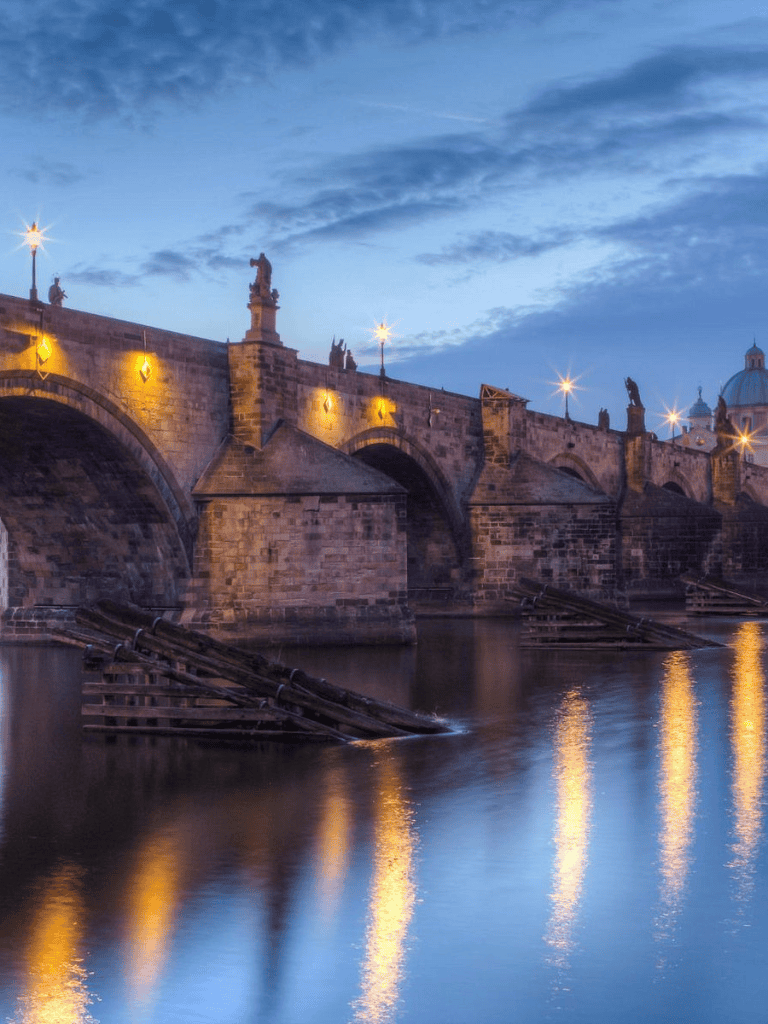 Ancient stone bridge over the river at dusk with illuminated statues and historic architecture.