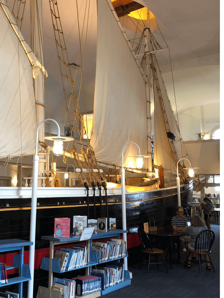 Sailboat exhibit in a library surrounded by books and reading areas, showcasing maritime history and navigation.