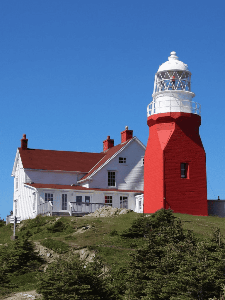 Lighthouse on rocky hill with white house, blue sky, and greenery, iconic coastal beacon.