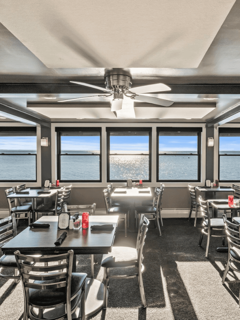 Sunlit waterfront restaurant dining area with ocean views, modern decor, and ceiling fan.