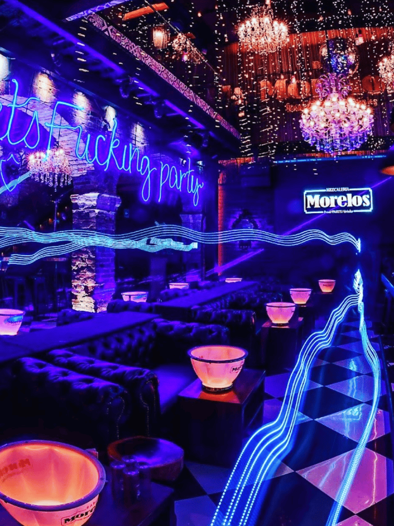 Neon "Fucking party" sign with chandeliers and vibrant lighting at a lively nightclub.
