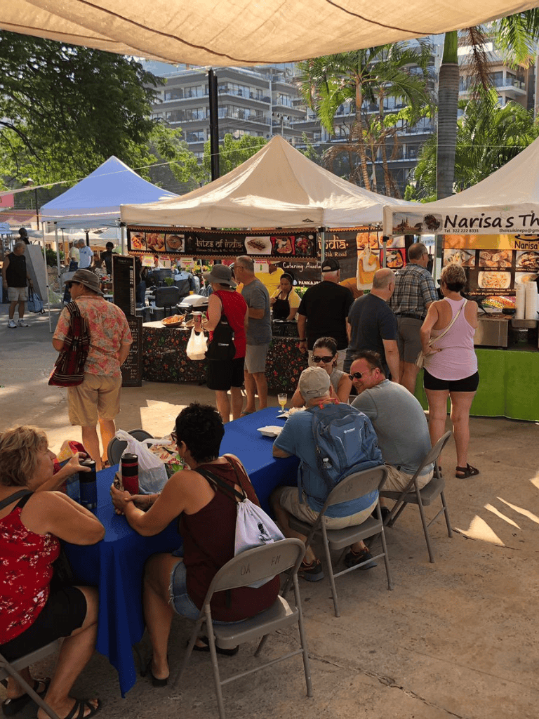 Colorful outdoor food market with people enjoying meals and browsing vendor stalls under tents.