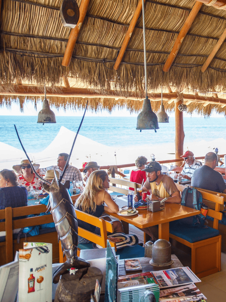 Colorful beachfront restaurant with thatched roof, ocean view, and lively diners enjoying meals.