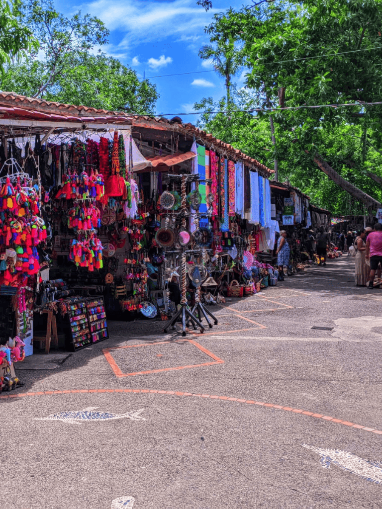 Colorful street market stalls with handmade crafts and souvenirs, lush green trees, and vibrant outdoor shopping scene.