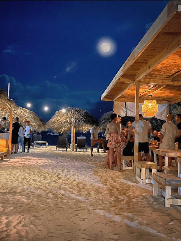Moonlit beach bar with tropical thatched umbrellas and lively group of people enjoying nighttime.