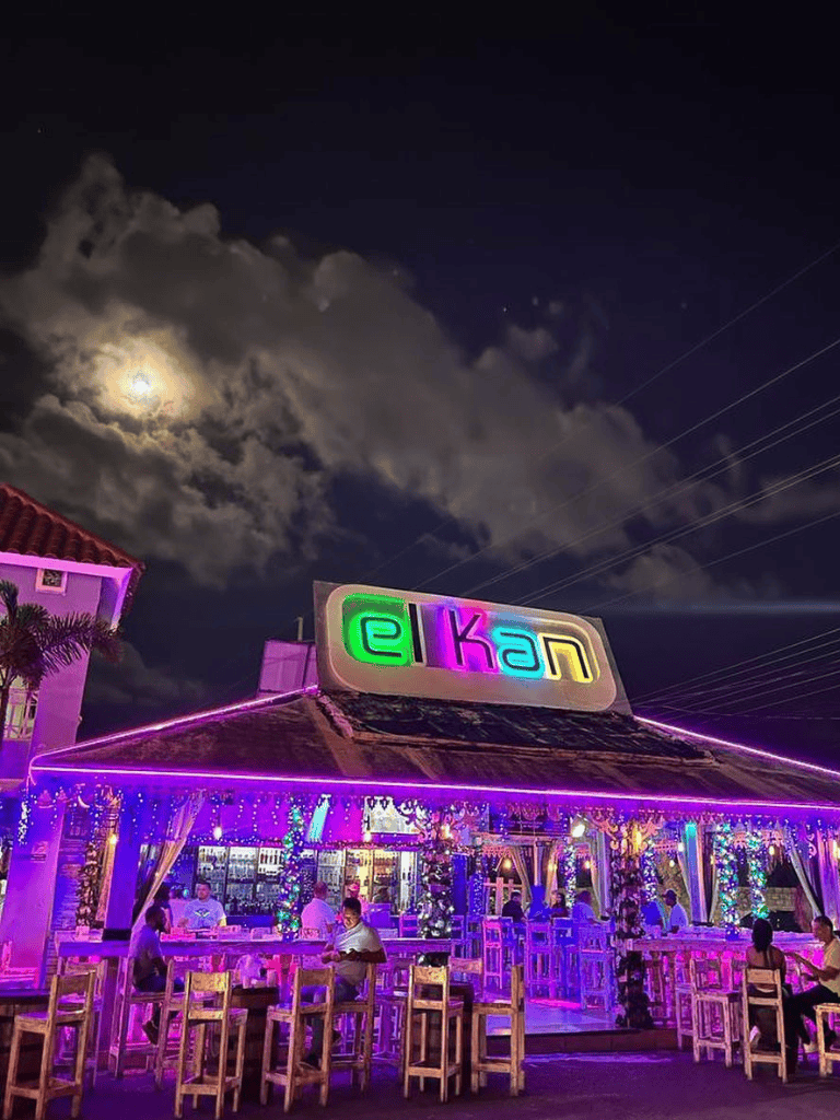 Colorful neon-lit restaurant at night with outdoor seating and illuminated sign "elkan" under cloudy moonlit sky.