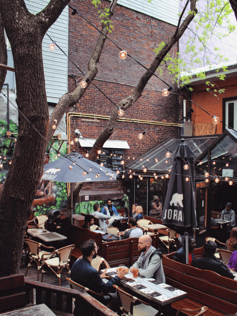 Outdoor restaurant patio with string lights and group of people enjoying drinks and conversation Relaxed outdoor dining scene with string lights, trees, and social atmosphere in urban setting.