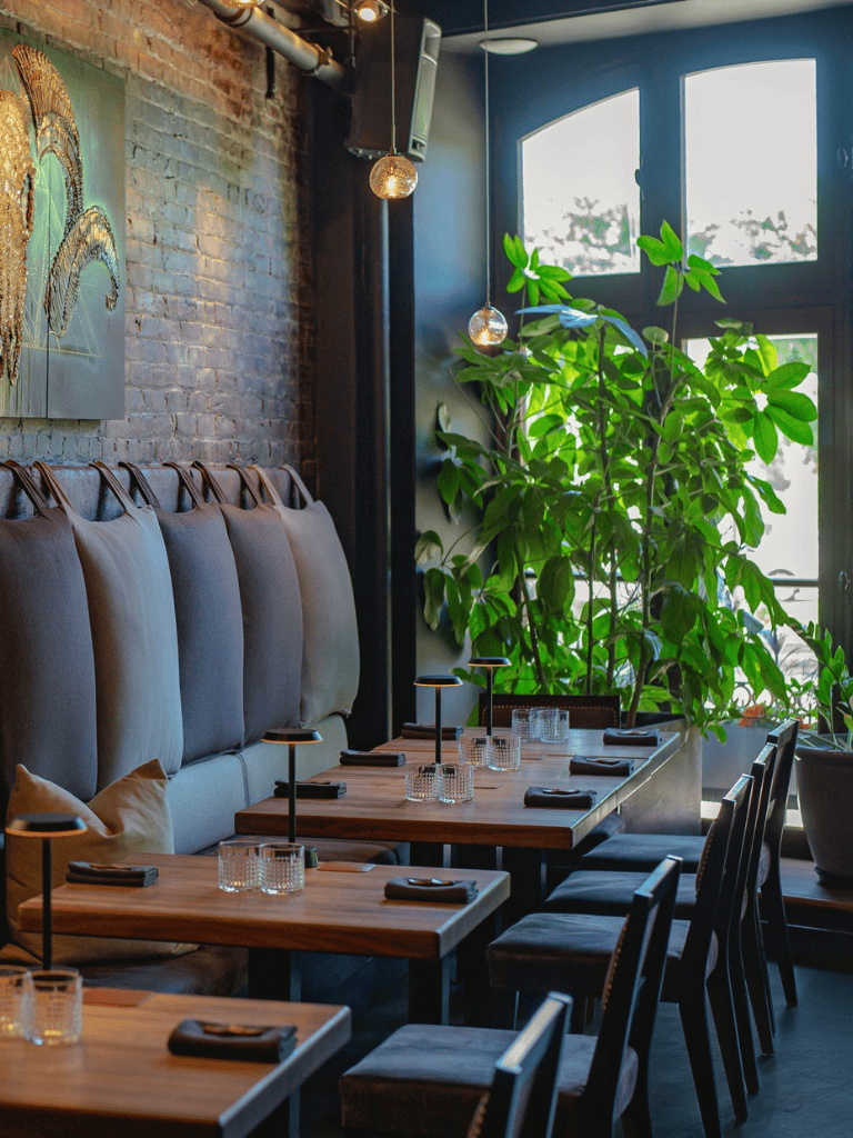 Bright restaurant interior with cozy seating, large green plants, and natural light from arched windows.
