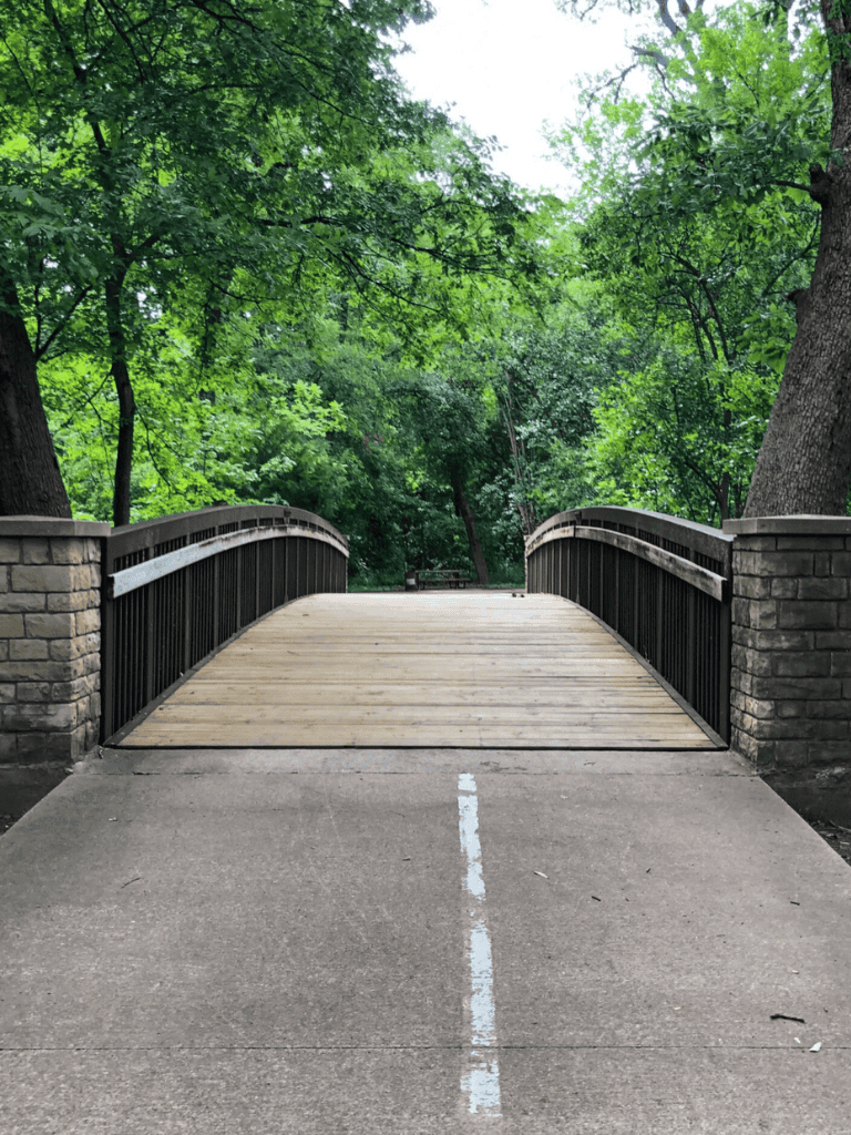 Wooden park bridge in a lush green forest with safety railings and brick supports, ideal for outdoor walking and nature exploration.