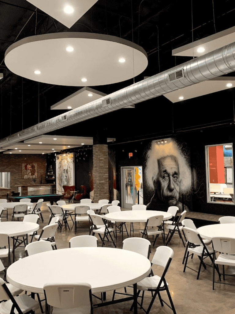 Modern restaurant interior with round tables, white chairs, and artistic portraits on brick walls.