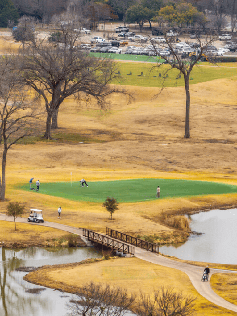 Golf course with players, trees, water features, and parking lot in the background.