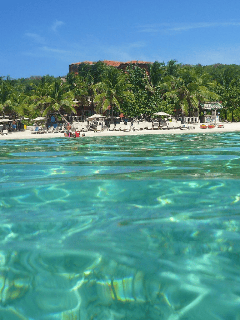 Turquoise Caribbean water with a resort and palm trees on sandy beach under blue sky.