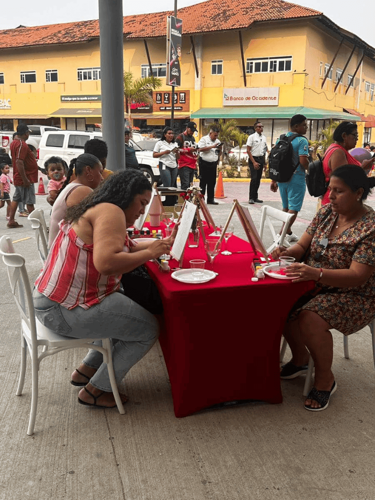 Colorful outdoor painting event in a vibrant city street, with participants creating artwork at a table.