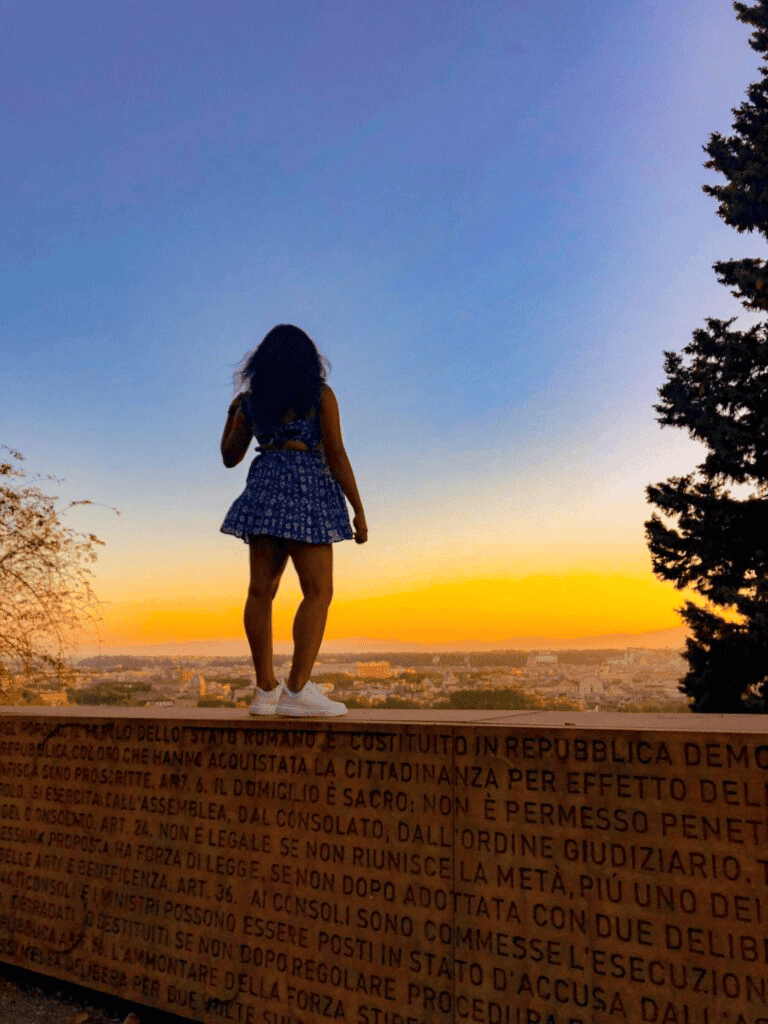 A girl standing on a ledge overlooking a city at sunset with trees nearby.