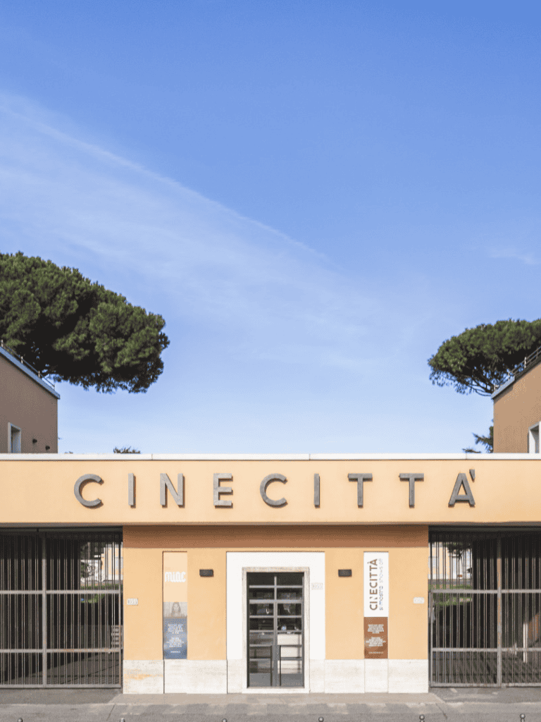 Modern Cinecittà building in Italy with clear signage and gated entrance, surrounded by trees, under a bright blue sky.