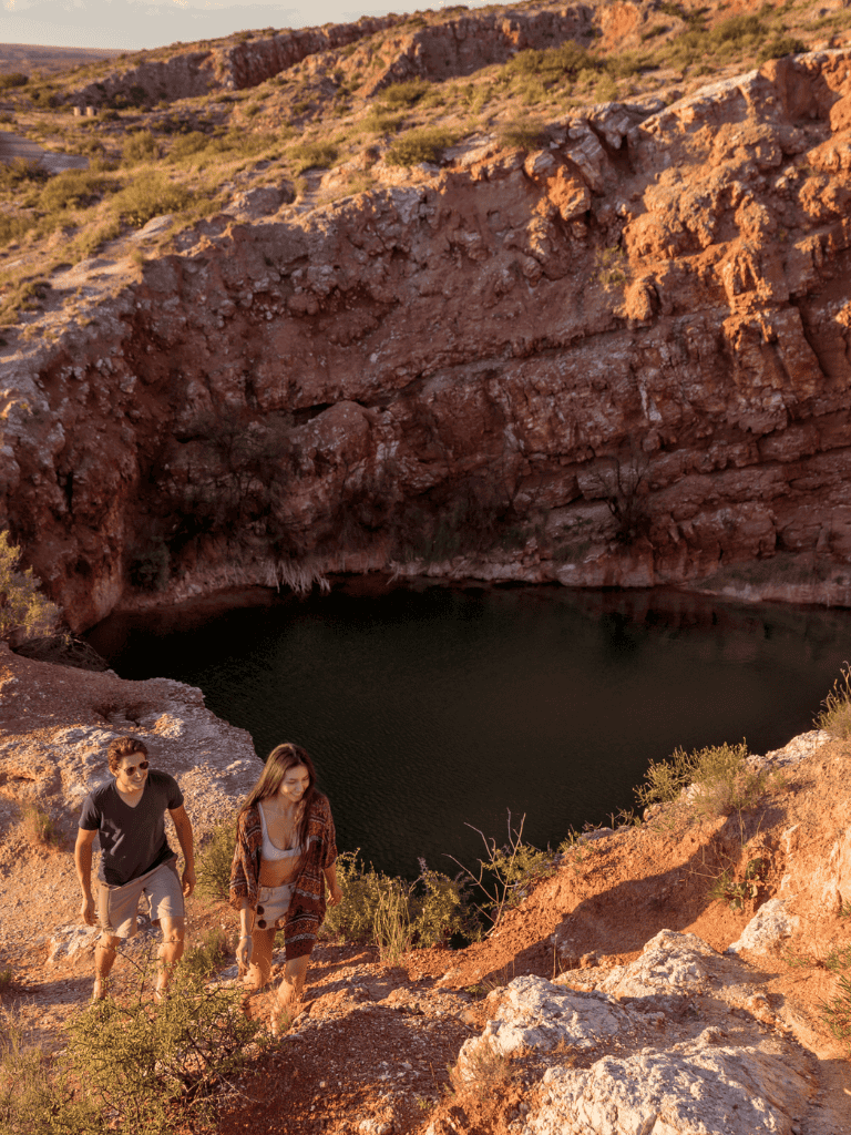 Sunset hike at a desert crater with two people exploring rugged terrain near a water-filled volcanic caldera.