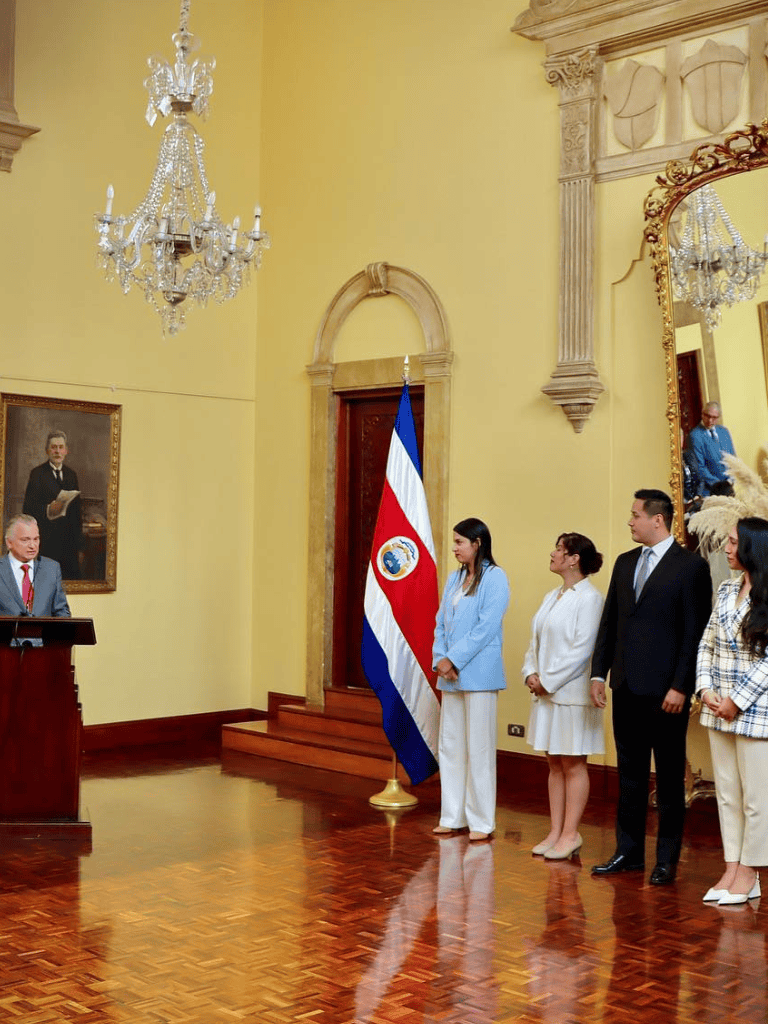 Costa Rican flag raising ceremony in an elegant historic room with chandeliers and a portrait on the wall.