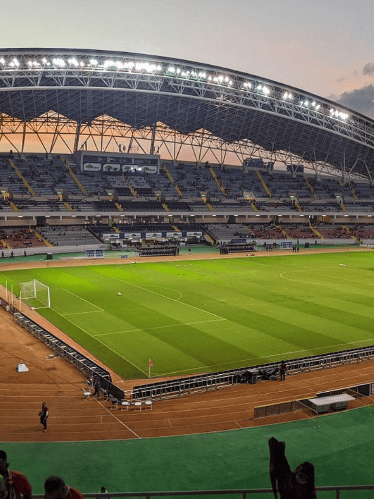 Bright stadium with green soccer field during sunset, seating, and track surrounding the field.