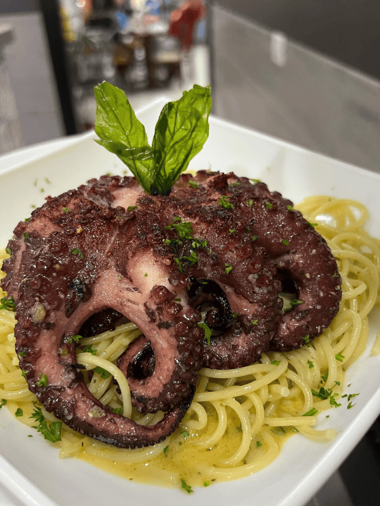 Grilled octopus over spaghetti with herbs, served in a white dish, delicious seafood pasta dish.