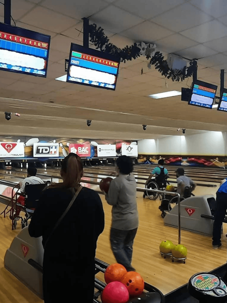 Brightly lit indoor bowling alley with players aiming and rolling balls, digital scoreboards overhead, and vibrant bowling balls on racks.