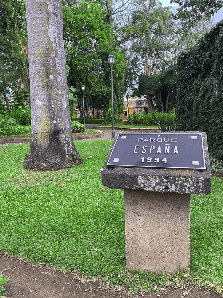 Lush green park with a historical plaque for Parque España, established in 1994, surrounded by trees and vintage lampposts.
