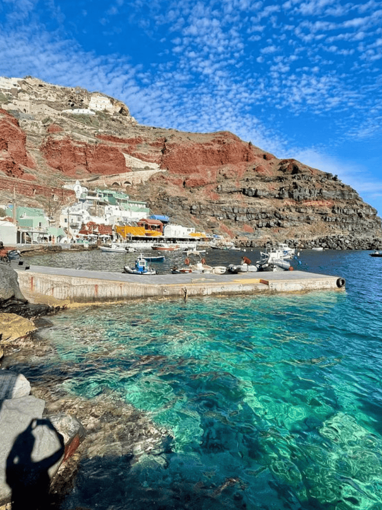Colorful coastal village with red cliffs and boats in crystal-clear water, scenic seaside destination in Greece.