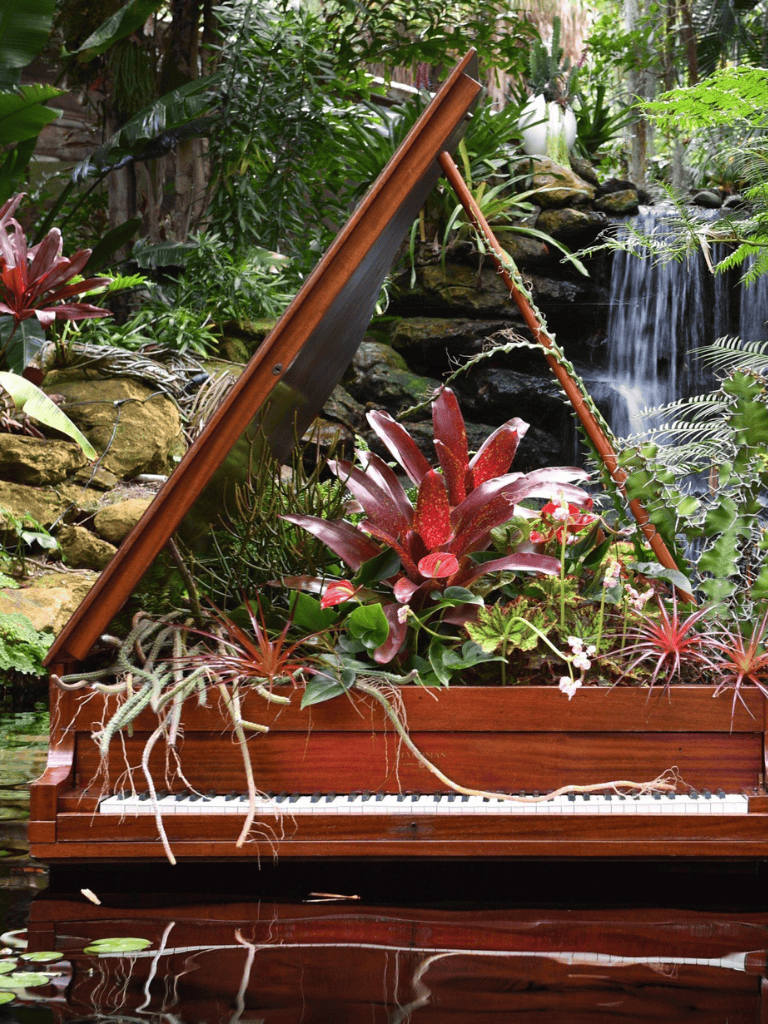 Lush tropical garden with a grand piano adorned with vibrant plants and water feature scene.
