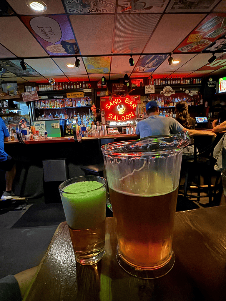 Cold brews and cocktails at a vibrant bar interior with neon "Sand Bar Saloon" sign, cozy atmosphere, and bar patrons.