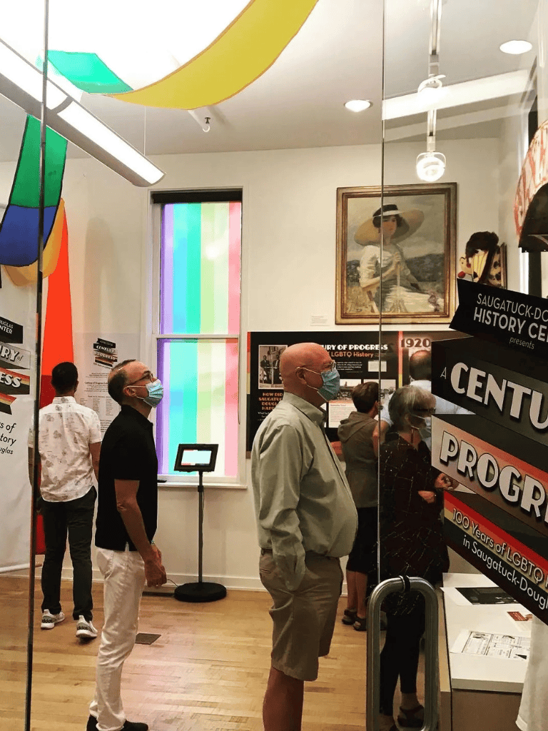 LGBTQ+ history exhibit at Saugatuck-Douglas History Center with visitors wearing masks and viewing informative displays.