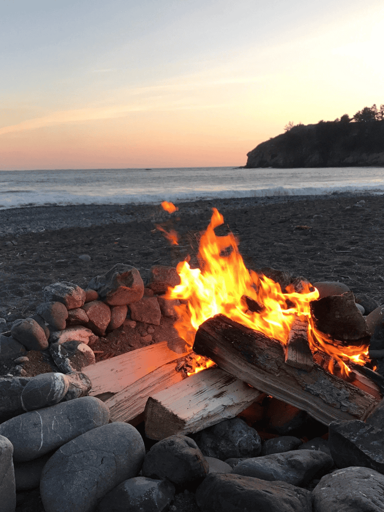Cozy beach campfire overlooking ocean at sunset, relaxing coastal outdoor fire pit scene.