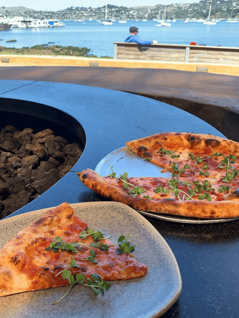Delicious pizza slices with fresh herbs at outdoor waterfront dining with scenic views.