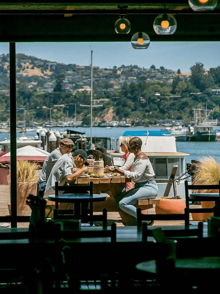 Relaxed people dining by the waterfront at a cozy outdoor cafe with boats and scenic hills in the background.