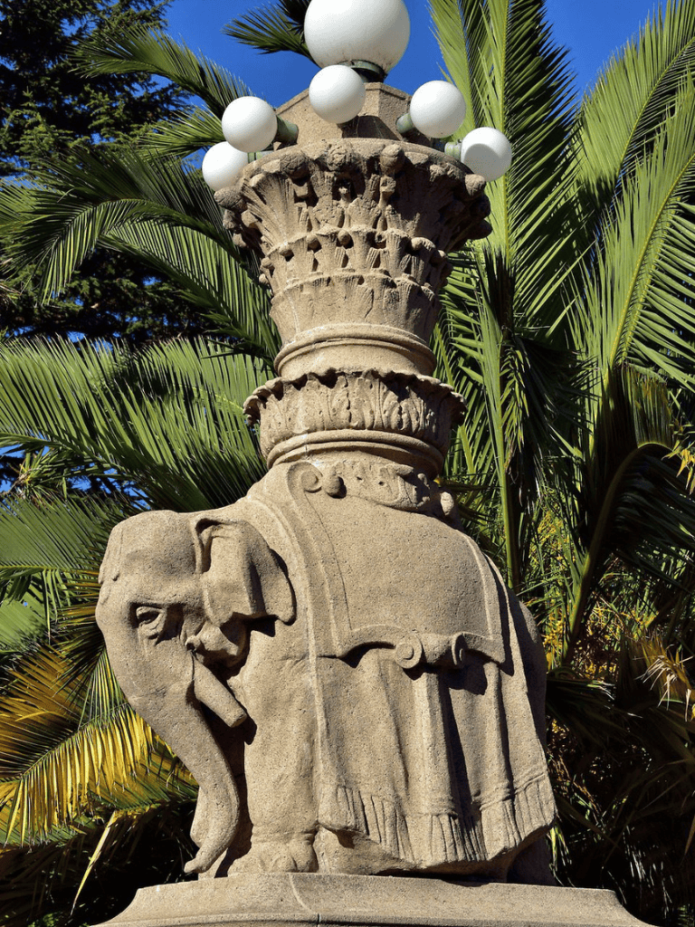 Elaborate stone street lamp with elephant detail, surrounded by lush palm trees and blue sky, representing historic architecture and tropical scenery.