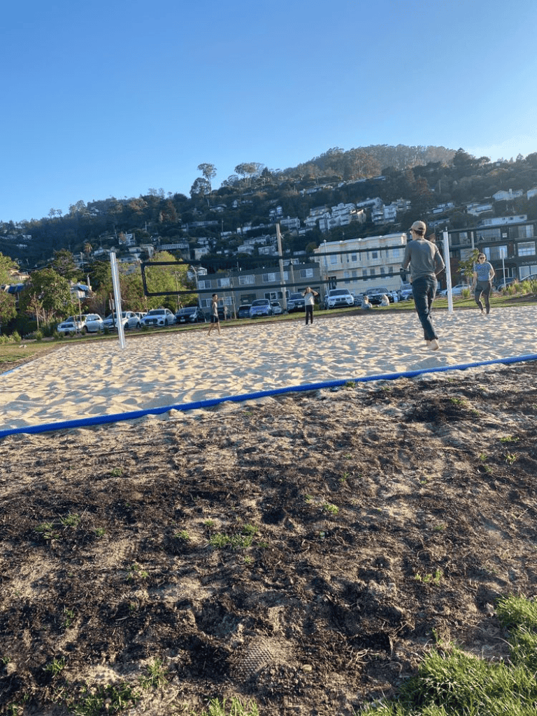 Cloudy sky and hillside neighborhood surrounding a beach volleyball court with players and spectators.