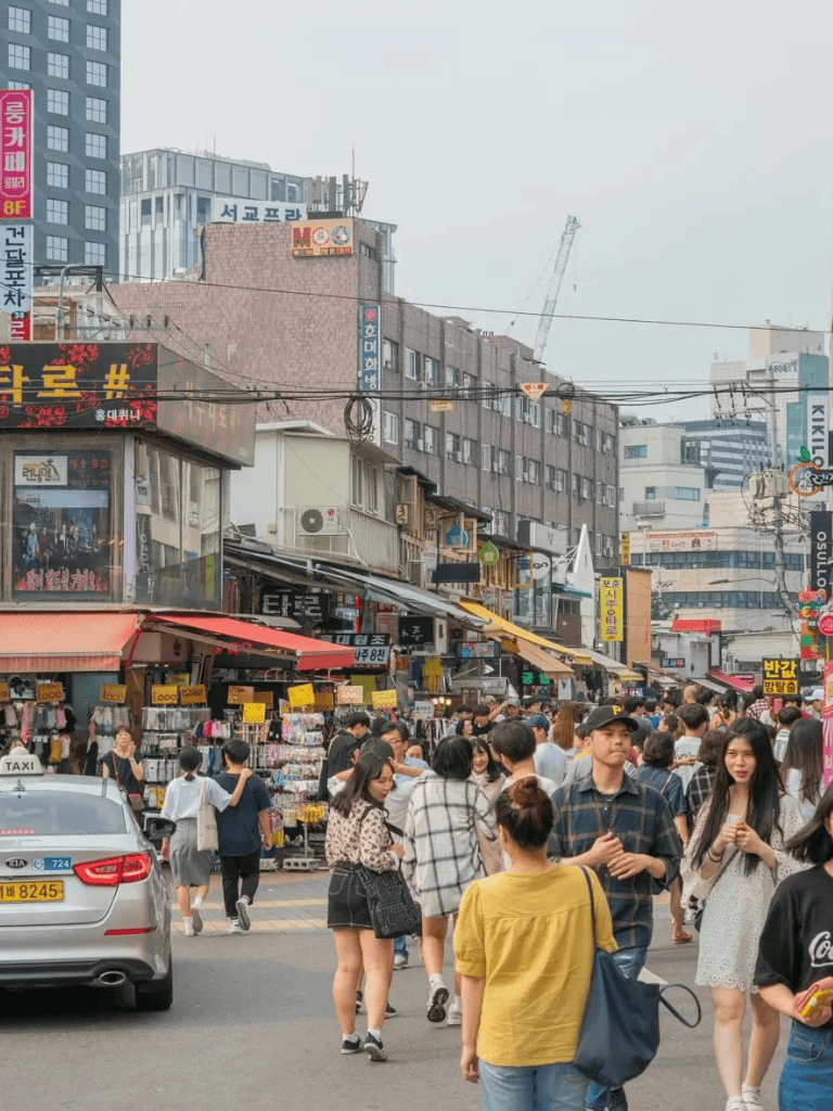 Busy street market in Korea with shoppers and storefronts, vibrant urban shopping district, street vendors, and cityscape.