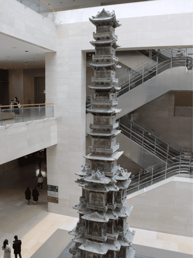 Intricate wooden pagoda replica displayed inside modern museum with escalators in background.