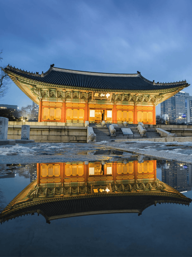 Traditional Korean temple illuminated at dusk with city skyline in background.