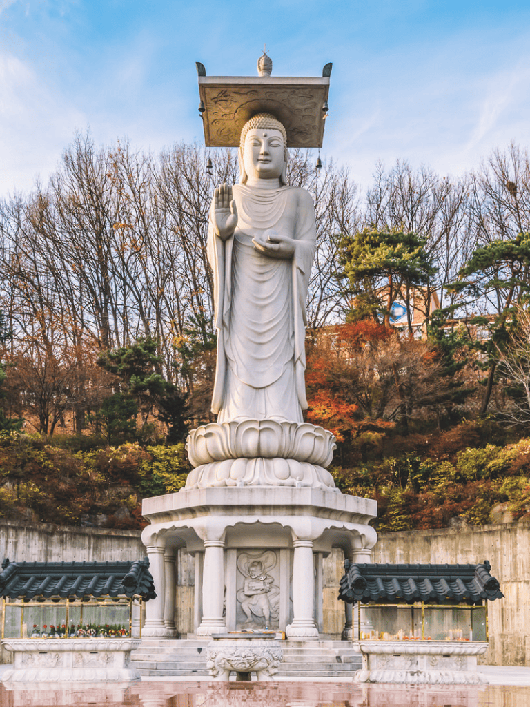Serene white Buddha statue in a peaceful outdoor garden, surrounded by autumn foliage and traditional Asian architecture.